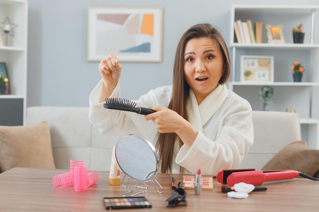 Giovane donna asiatica con i capelli lunghi scuri che si siede al tavolo da toeletta a casa facendo la routine di trucco mattutina che sembra confusa pulendo la spazzola dai capelli che soffrono di caduta dei capelli