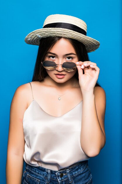 Giovane donna asiatica con la posa di sorpresa isolata su fondo blu. Ritratto di bella donna asiatica in cappello di paglia e occhiali da sole su sfondo blu