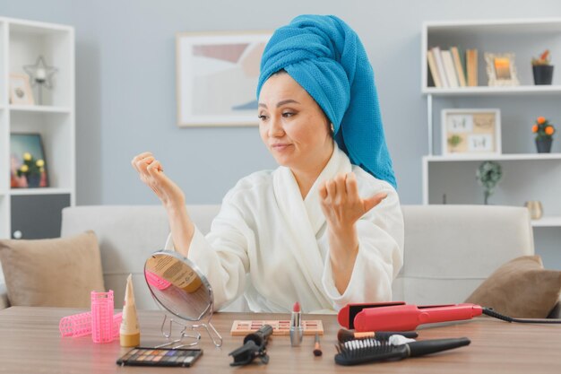 giovane donna asiatica con un asciugamano in testa seduta al tavolo da toeletta a casa guardando le unghie con espressione scettica facendo routine di trucco mattutino