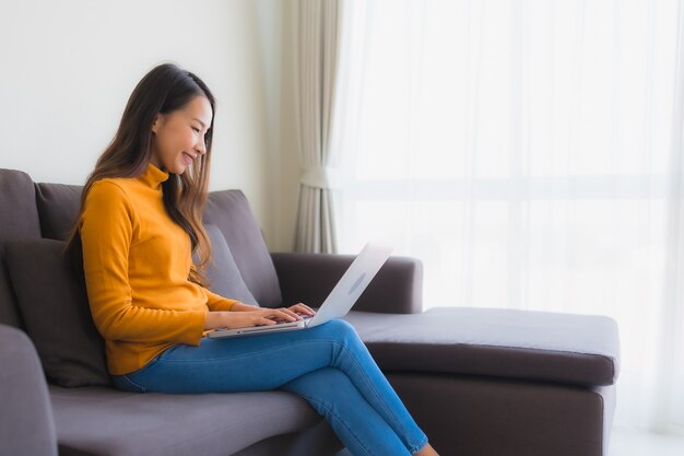 Giovane donna asiatica del ritratto che per mezzo del taccuino del computer portatile sul sofà