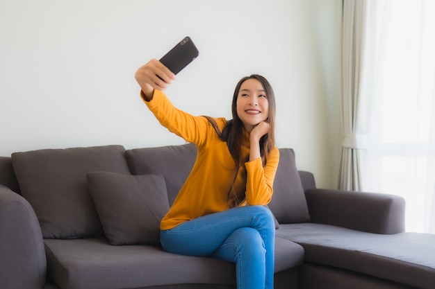Giovane donna asiatica del ritratto che utilizza telefono cellulare astuto sul sofà con il cuscino nel salone