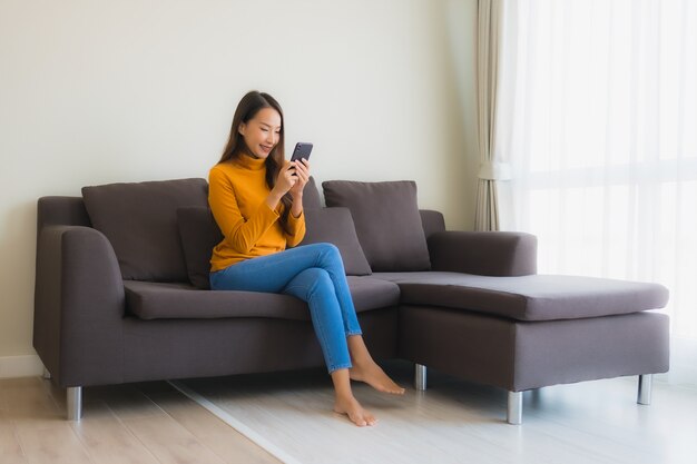 Giovane donna asiatica del ritratto che utilizza telefono cellulare astuto sul sofà con il cuscino nel salone