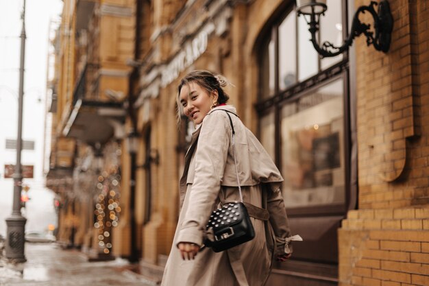 Giovane donna asiatica di buon umore cammina per la città in elegante trench con piccola borsa nera