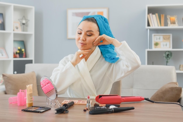 Giovane donna asiatica felice con l'asciugamano in testa seduto al tavolo da toeletta a casa interiore guardando nello specchio facendo massaggio facciale bellezza e concetto di cosmetici per il viso