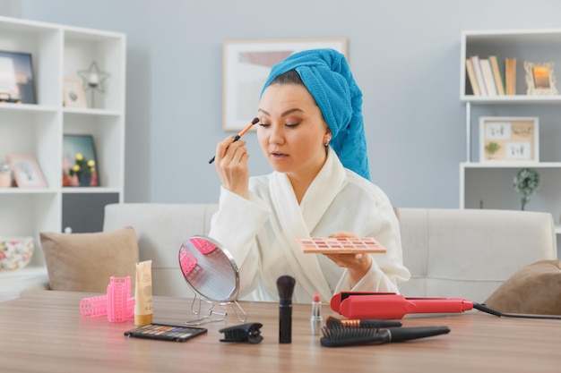 Giovane donna asiatica felice con l'asciugamano in testa seduto al tavolo da toeletta a casa interni applicando ombretti guardando lo specchio facendo trucco mattutino bellezza di routine e concetto di cosmetici per il viso