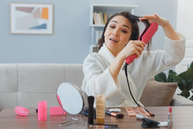 Giovane donna asiatica felice con un asciugamano in accappatoio seduto al tavolo da toeletta a casa ferro arricciacapelli interno che fa styling facendo routine di trucco mattutino