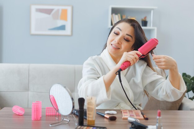 Giovane donna asiatica felice con un asciugamano in accappatoio seduto al tavolo da toeletta all'interno di casa utilizzando ferro arricciacapelli facendo styling facendo routine di trucco mattutino