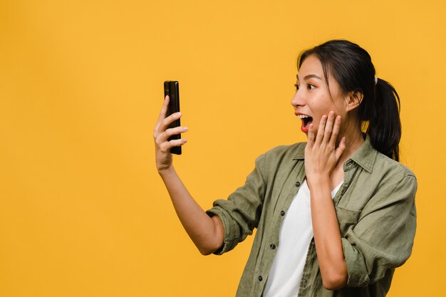 Giovane donna asiatica sorpresa che usa il telefono cellulare con espressione positiva, sorride ampiamente, vestita con abiti casual e sta isolata sul muro giallo. La donna felice adorabile felice si rallegra del successo.