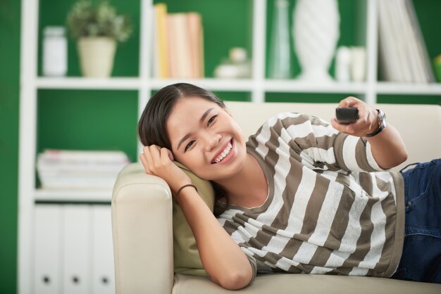 Giovane donna asiatica sorridente che si trova sullo strato a casa, guardando diritto e premendo sul telecomando