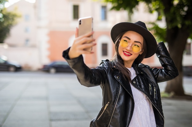 Giovane donna asiatica sorridente in occhiali da sole che prendono selfie sulla via della città
