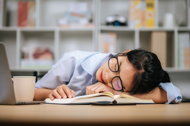 Giovane donna asiatica stanca in occhiali che dorme sulla scrivania con laptop e libro di testo a casa