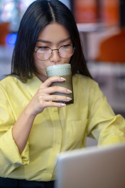 Giovane donna asiatica sveglia in occhiali al computer portatile che mangia caffè