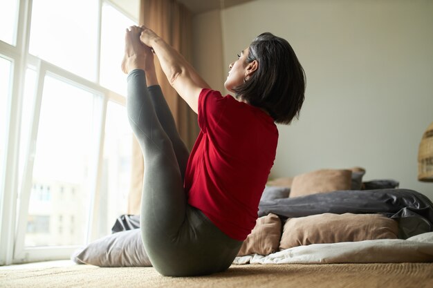 Giovane donna atletica con corpo in forma forte, praticare yoga in camera da letto, seduto rivolto verso l'alto intenso tratto posa