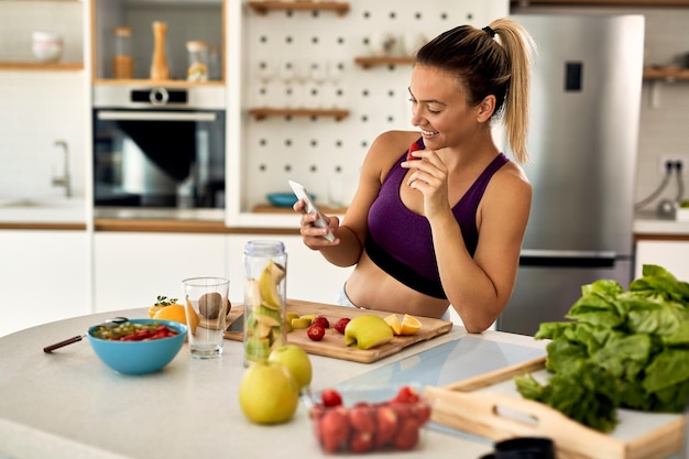 Giovane donna atletica felice che mangia frutta mentre messaggi di testo sul telefono cellulare in cucina