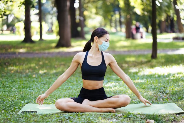 Giovane donna atletica in una maschera protettiva medica, facendo yoga nel parco al mattino, formazione delle donne su una stuoia di yoga