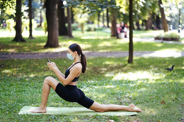 Giovane donna atletica in una maschera protettiva medica, facendo yoga nel parco