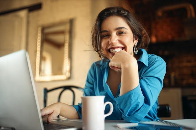 Giovane donna attraente allegra che gode del lavoro distante, seduto alla scrivania utilizzando il computer portatile, bevendo caffè. Bella donna blogger che lavora da casa, carica video sul suo canale, sorridendo