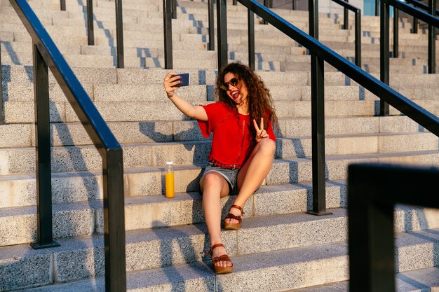 Giovane donna attraente allegra che mostra un gesto di pace durante la presa del selfie