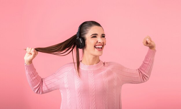 Giovane donna attraente che ascolta la musica in cuffie senza fili che indossano maglione rosa sorridente felice stato d'animo positivo in posa su sfondo rosa isolato divertente espressione emotiva del viso