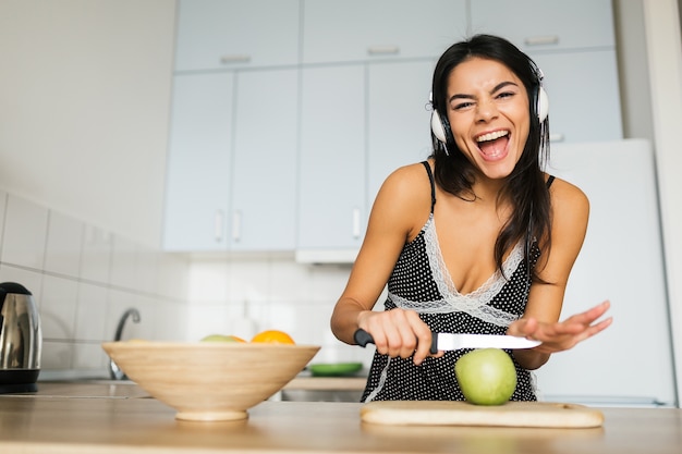 Giovane donna attraente che cucina in cucina al mattino, fare insalata di frutta, tagliare la mela con il coltello, sorridente, felice, positivo, stile di vita sano, ascoltare musica in cuffia, ridere, divertirsi