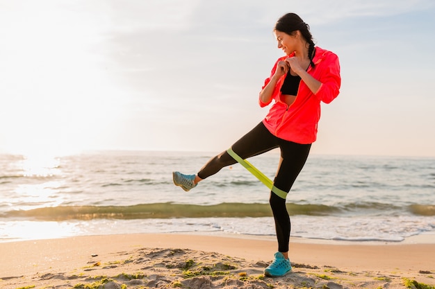 Giovane donna attraente che fa esercizi sportivi all'alba mattutina sulla spiaggia del mare, stile di vita sano, ascolto di musica sugli auricolari, indossa una giacca a vento rosa, facendo stretching in elastico