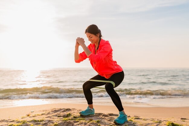 Giovane donna attraente che fa esercizi sportivi all'alba mattutina sulla spiaggia del mare, stile di vita sano, ascolto di musica sugli auricolari, indossa una giacca a vento rosa, facendo stretching in elastico