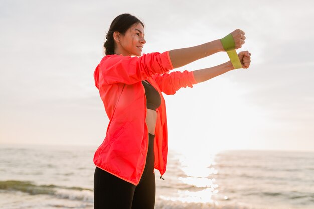 Giovane donna attraente che fa esercizi sportivi all'alba mattutina sulla spiaggia del mare, stile di vita sano, ascolto di musica sugli auricolari, indossa una giacca a vento rosa, facendo stretching in elastico