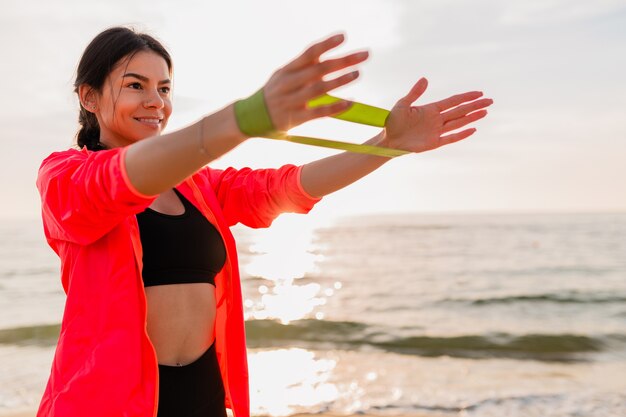 Giovane donna attraente che fa esercizi sportivi all'alba mattutina sulla spiaggia del mare, stile di vita sano, ascolto di musica sugli auricolari, indossa una giacca a vento rosa, facendo stretching in elastico