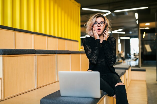 Giovane donna attraente che si siede nell'aula di conferenza che lavora al computer portatile che indossa i vetri, sala moderna, istruzione dello studente online, espressione scioccata del fronte