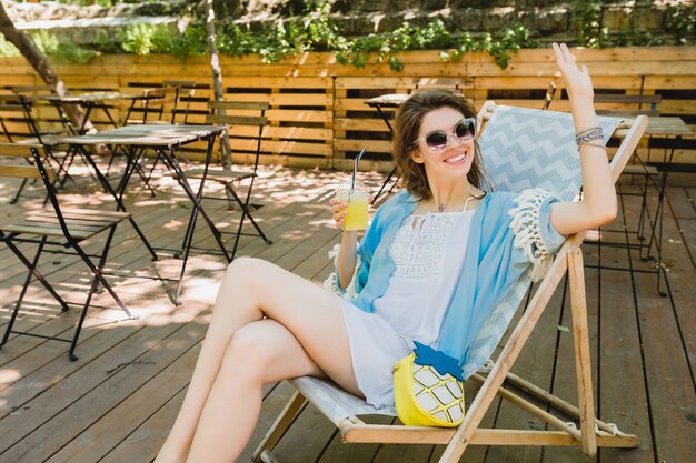 Giovane donna attraente che si siede nella sedia a sdraio in abito di moda estiva, abito bianco, mantello blu, occhiali da sole, sorridente, bere limonata, accessori alla moda, relax in vacanza