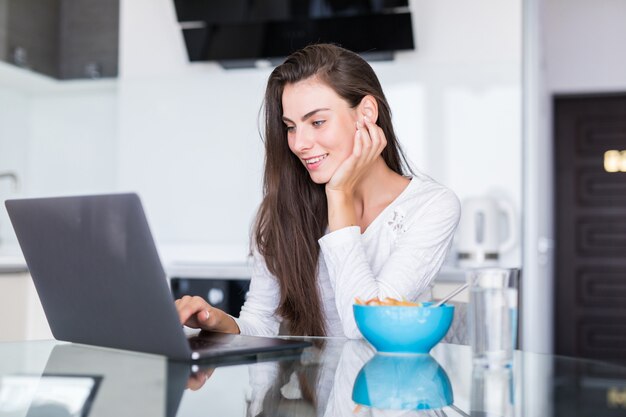 Giovane donna attraente che utilizza computer portatile a colazione e che si siede nella cucina.