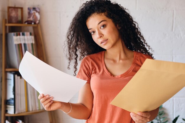 Giovane donna attraente con i capelli ricci scuri in maglietta che guarda sognante nella macchina fotografica tenendo la lettera con i risultati dell'esame nelle mani a casa accogliente