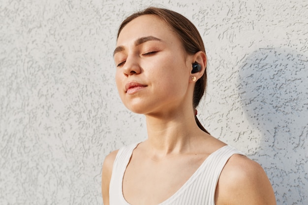 Giovane donna attraente con i capelli scuri che indossa un top bianco, ascolta musica, usa gli airpod, tiene gli occhi chiusi, si gode la canzone preferita durante l'allenamento all'aperto.