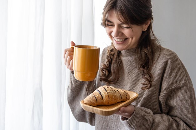 Giovane donna attraente con una tazza di bevanda calda e un croissant vicino alla finestra