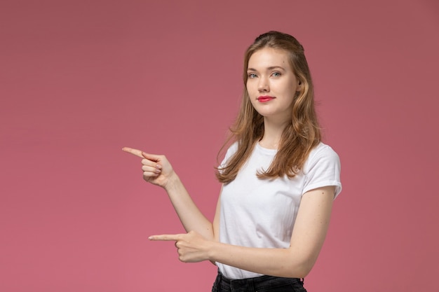 Giovane donna attraente di vista frontale in maglietta bianca e pantaloni neri che posano sulla foto a colori di posa femminile del modello della parete rosa