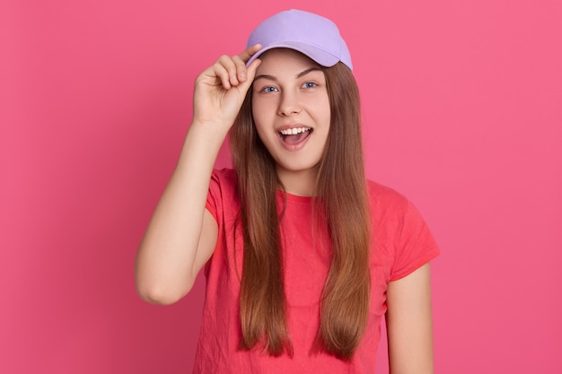 Giovane donna attraente eccitata che indossa abbigliamento casual e berretto da baseball, sembra felice, urlando qualcosa, toccando la visiera del cappello