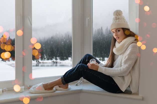 Giovane donna attraente in elegante maglione lavorato a maglia bianco, sciarpa e cappello seduto a casa sul davanzale della finestra a Natale che tiene la decorazione presente della palla di neve di vetro, vista della foresta di inverno, bokeh delle luci