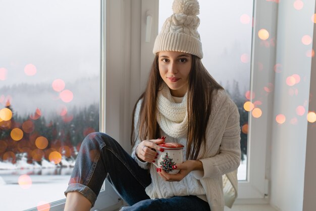 Giovane donna attraente in elegante maglione lavorato a maglia bianco, sciarpa e cappello seduto a casa sul davanzale della finestra a Natale che tiene tazza bere tè caldo, vista sullo sfondo foresta invernale, luci bokeh