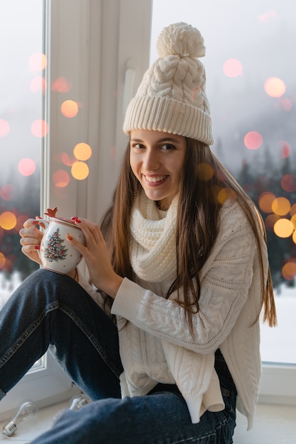 Giovane donna attraente in elegante maglione lavorato a maglia bianco, sciarpa e cappello seduto a casa sul davanzale della finestra a Natale che tiene tazza bere tè caldo, vista sullo sfondo foresta invernale, luci bokeh