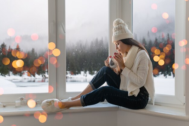 Giovane donna attraente in elegante maglione lavorato a maglia bianco, sciarpa e cappello seduto a casa sul davanzale della finestra a Natale che tiene tazza bere tè caldo, vista sullo sfondo foresta invernale, luci bokeh