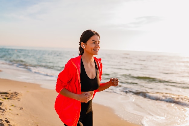 Giovane donna attraente sottile facendo esercizi sportivi all'alba mattutina a fare jogging sulla spiaggia del mare in abbigliamento sportivo, stile di vita sano, ascolto di musica sugli auricolari, indossa una giacca a vento rosa