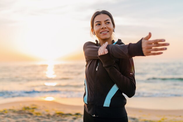 Giovane donna attraente sottile facendo esercizi sportivi sulla spiaggia di alba di mattina in abbigliamento sportivo, stile di vita sano, ascoltando musica con gli auricolari, facendo stretching per le braccia