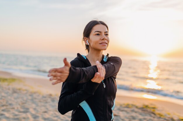 Giovane donna attraente sottile facendo esercizi sportivi sulla spiaggia di alba di mattina in abbigliamento sportivo, stile di vita sano, ascoltando musica con gli auricolari, facendo stretching per le mani