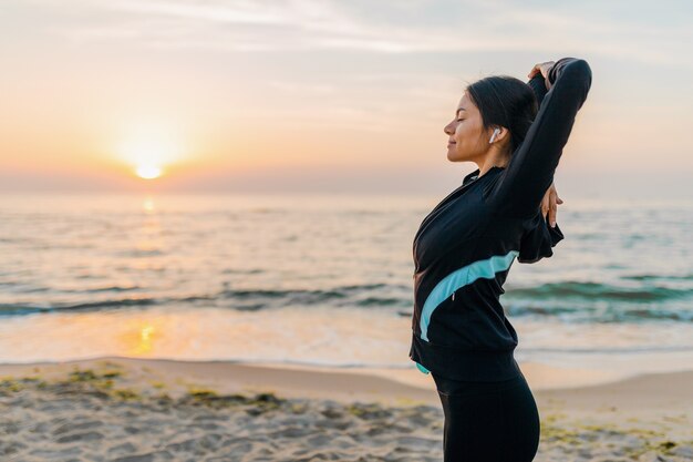 Giovane donna attraente sottile facendo esercizi sportivi sulla spiaggia di alba di mattina in abbigliamento sportivo, stile di vita sano, ascoltando musica con gli auricolari, facendo stretching