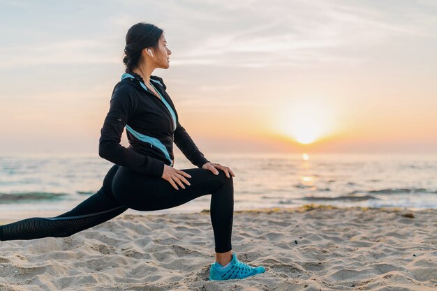 Giovane donna attraente sottile facendo esercizi sportivi sulla spiaggia di alba di mattina in abbigliamento sportivo, stile di vita sano, ascoltando musica con gli auricolari, facendo stretching