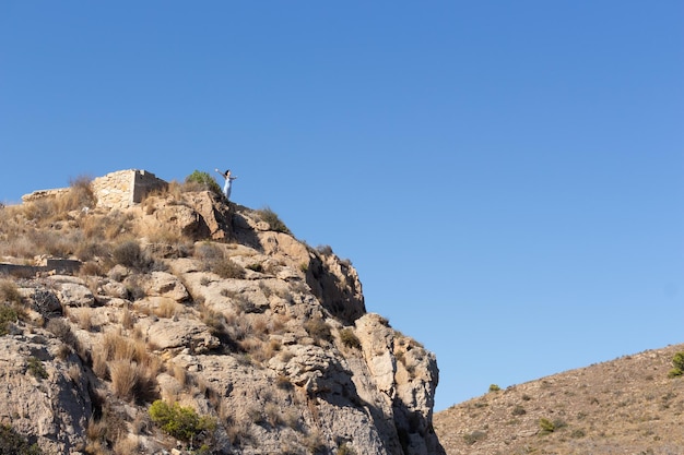 Giovane donna avventurosa in piedi su una scogliera rocciosa alzando le mani e ammirando il panorama