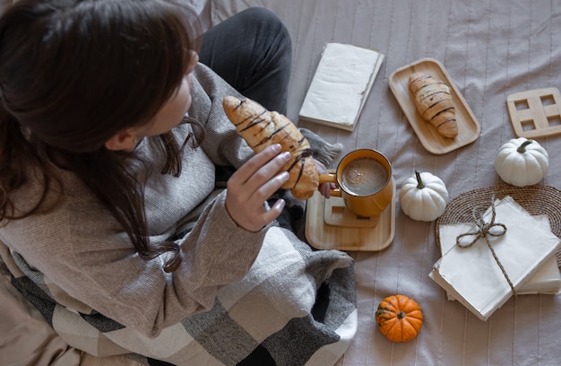 Giovane donna, avvolta in un plaid, che beve caffè con un croissant a letto, vista dall'alto, concetto autunnale.