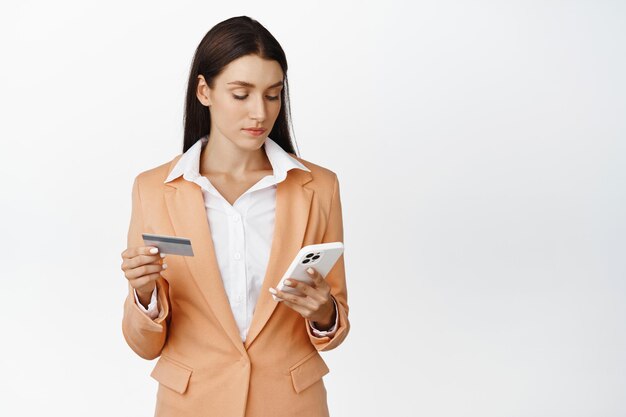 Giovane donna aziendale che paga sul telefono cellulare utilizzando la carta di credito e l'app per smartphone per ordinare effettuare acquisti online in piedi su sfondo bianco