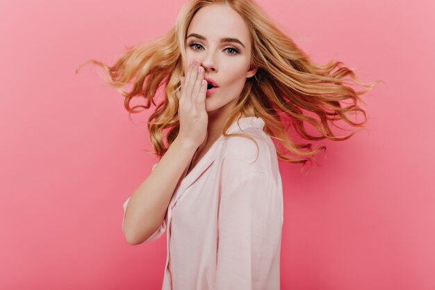 Giovane donna bianca vaga con capelli ricci che fluttuano in posa sulla parete rosa. Foto dell'interno della signora caucasica sorpresa in abiti da notte che copre la bocca con la mano.