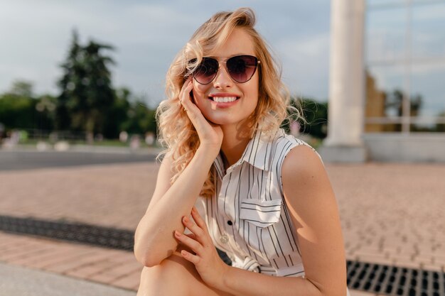 Giovane donna bionda alla moda carina attraente attraente che si siede nella via della città in vestito da cotone bianco di stile di modo di estate che indossa gli occhiali da sole